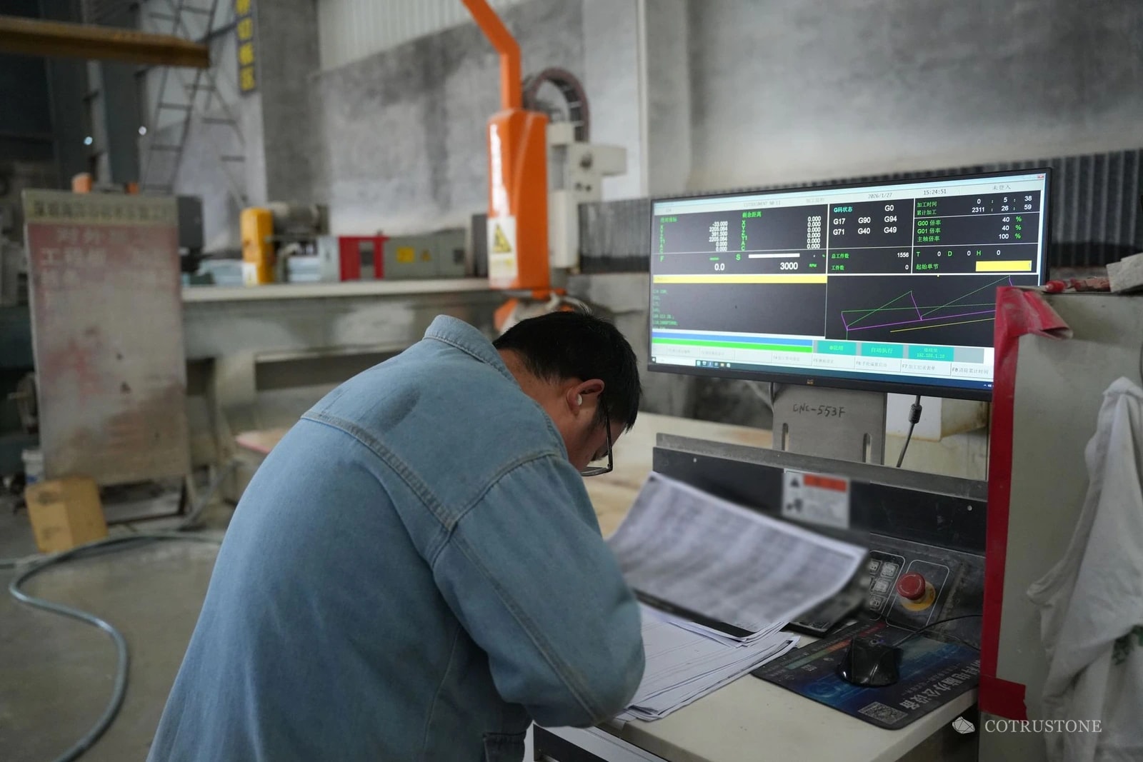 Operator reviewing production paperwork beside a stone machining control screen